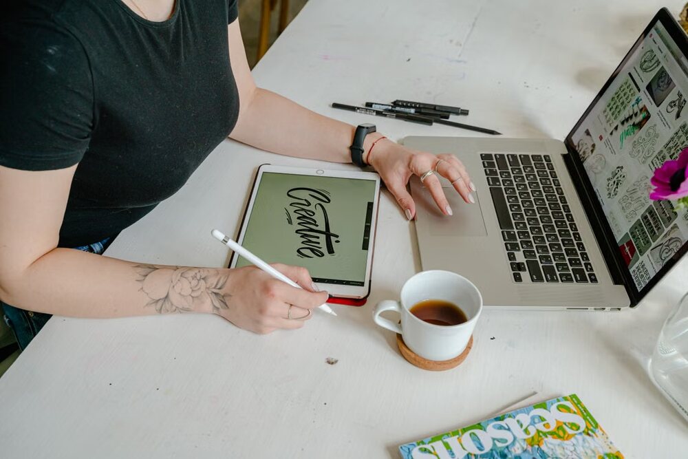Graphic designer creating digital lettering on a tablet beside a laptop.