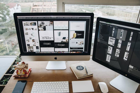 Dual-monitor Mac workspace showing web design layouts and creative software on a wooden desk.