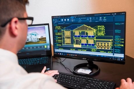 Man viewing a house elevation drawing on a monitor beside a laptop displaying a photo of a house
