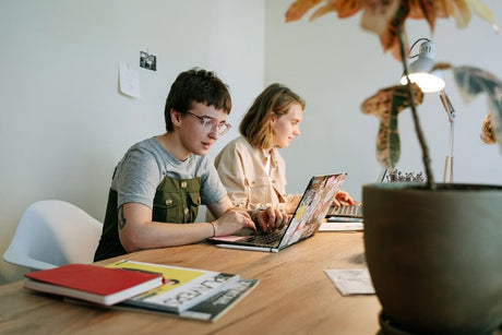 Two students working on laptops at a wooden table with notebooks and a potted plant in the foreground